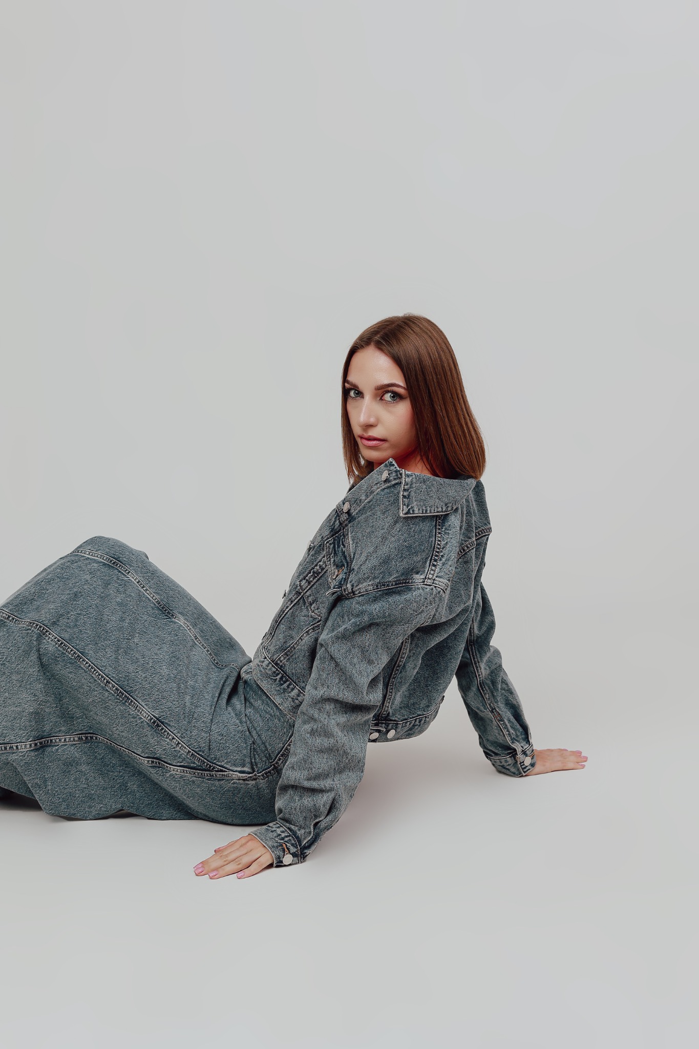 Kobieta w dżinsowym total looku, siedząca na podłodze, spogląda przez ramię w obiektyw, na neutralnym tle.