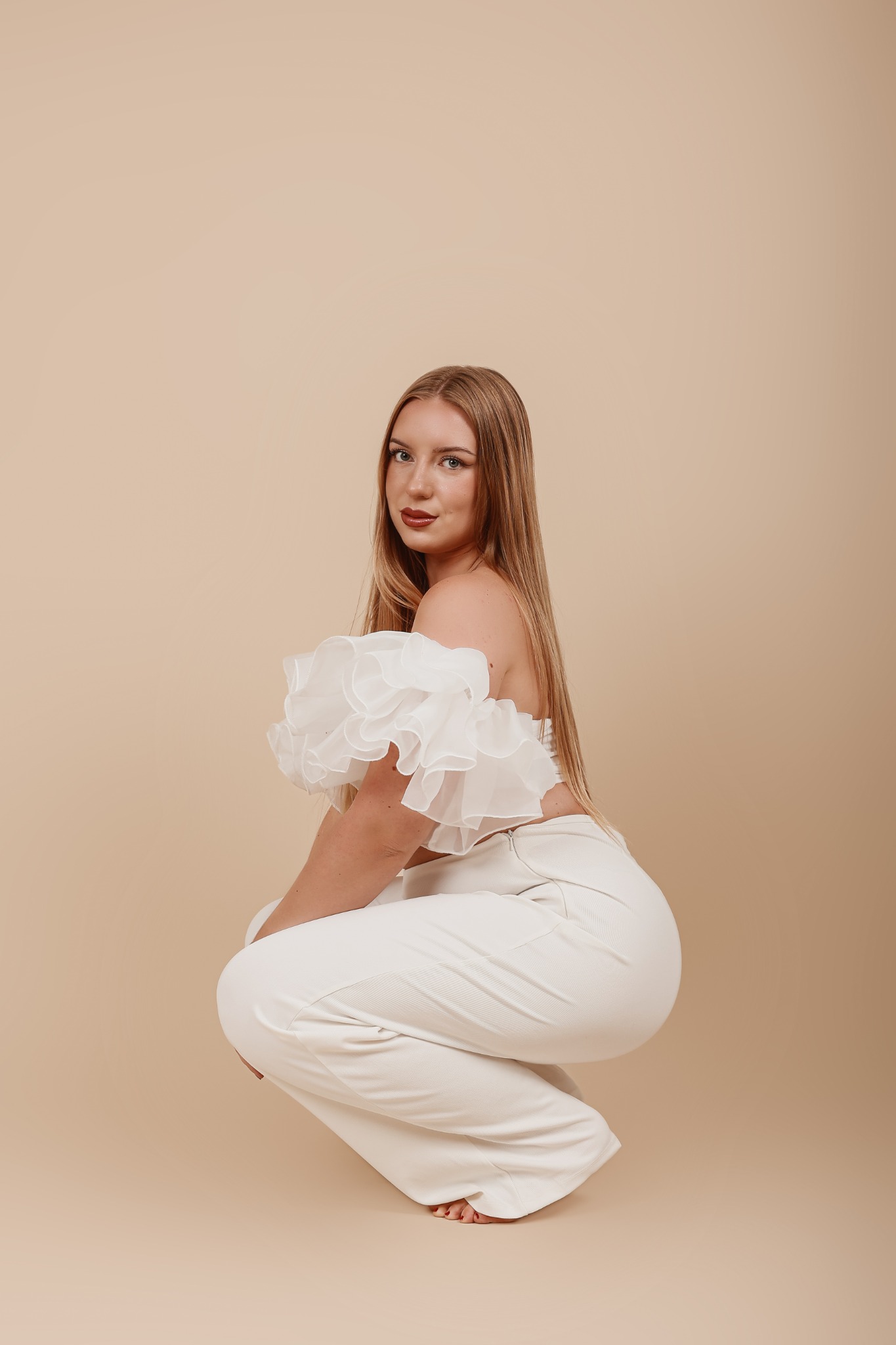 Portret kobiety w białym stroju, kucającej na beżowym tle, z bluzką z falbanami i długimi, prostymi włosami.