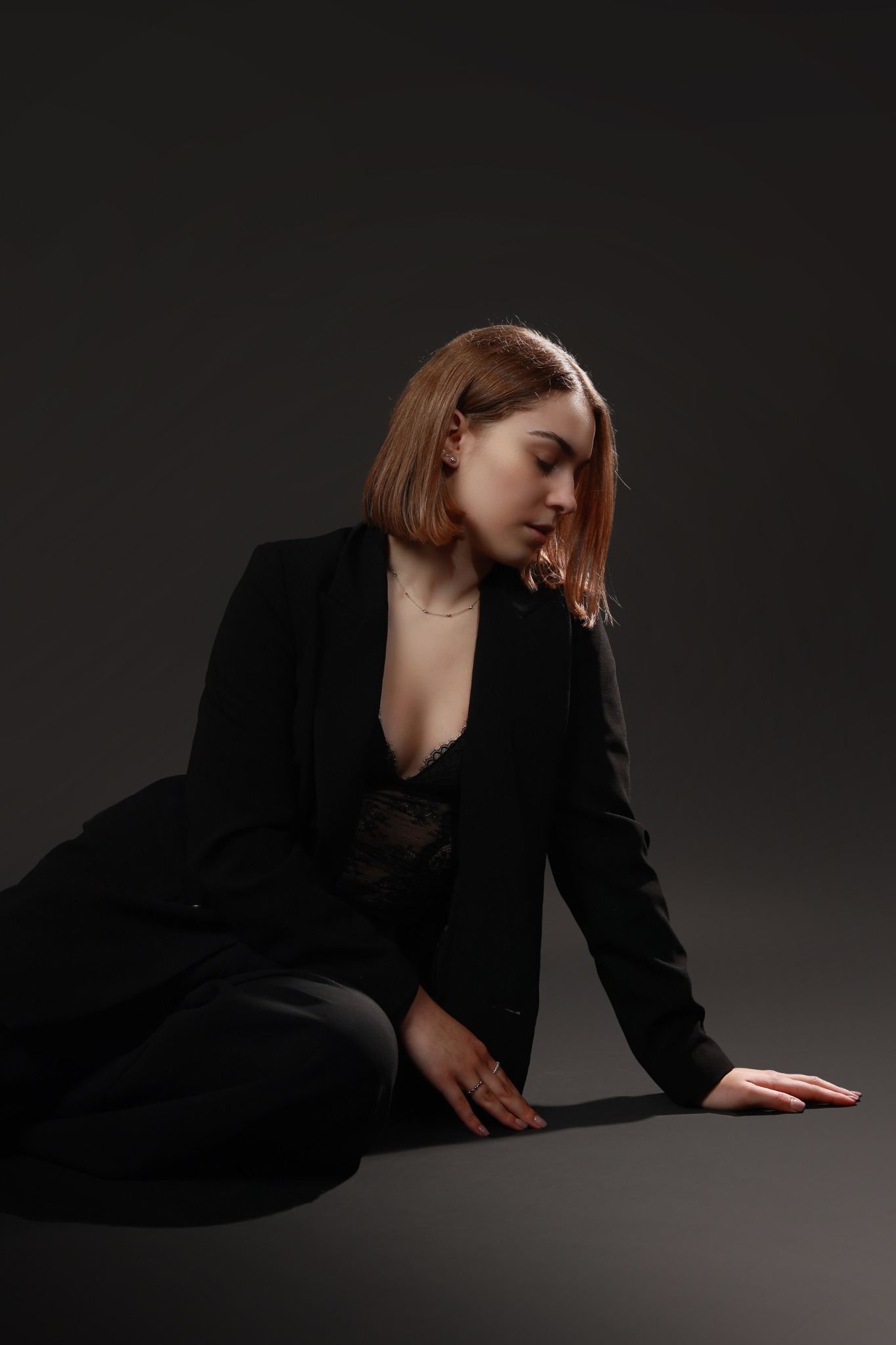 Portret kobiety w czarnym garniturze i koronkowej bieliźnie, siedzącej na szarym tle, z delikatnym oświetleniem podkreślającym jej rysy twarzy i subtelny urok