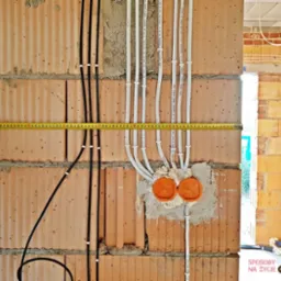 Surowe ściany z pustaków ceramicznych z poprowadzonymi przewodami elektrycznymi w białych i czarnych rurkach, pomiar odległości żółtą miarką zwijaną, widoczne pomarańczowe puszki elektryczne zalane...
