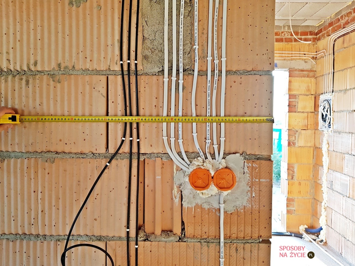 Surowe ściany z pustaków ceramicznych z poprowadzonymi przewodami elektrycznymi w białych i czarnych rurkach, pomiar odległości żółtą miarką zwijaną, widoczne pomarańczowe puszki elektryczne zalane...