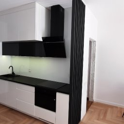 SEBMEBEL SEBASTIAN KWARCIANY - Nowoczesna kuchnia z białymi, lakierowanymi szafkami, czarnym blatem i okapem, widoczny fragment jadalni ze szklanym stołem i owocami.