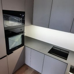 SEBMEBEL SEBASTIAN KWARCIANY - Nowoczesna kuchnia z szarymi frontami szafek, wbudowanym piekarnikiem Samsung w folii ochronnej i drewnianą podłogą.