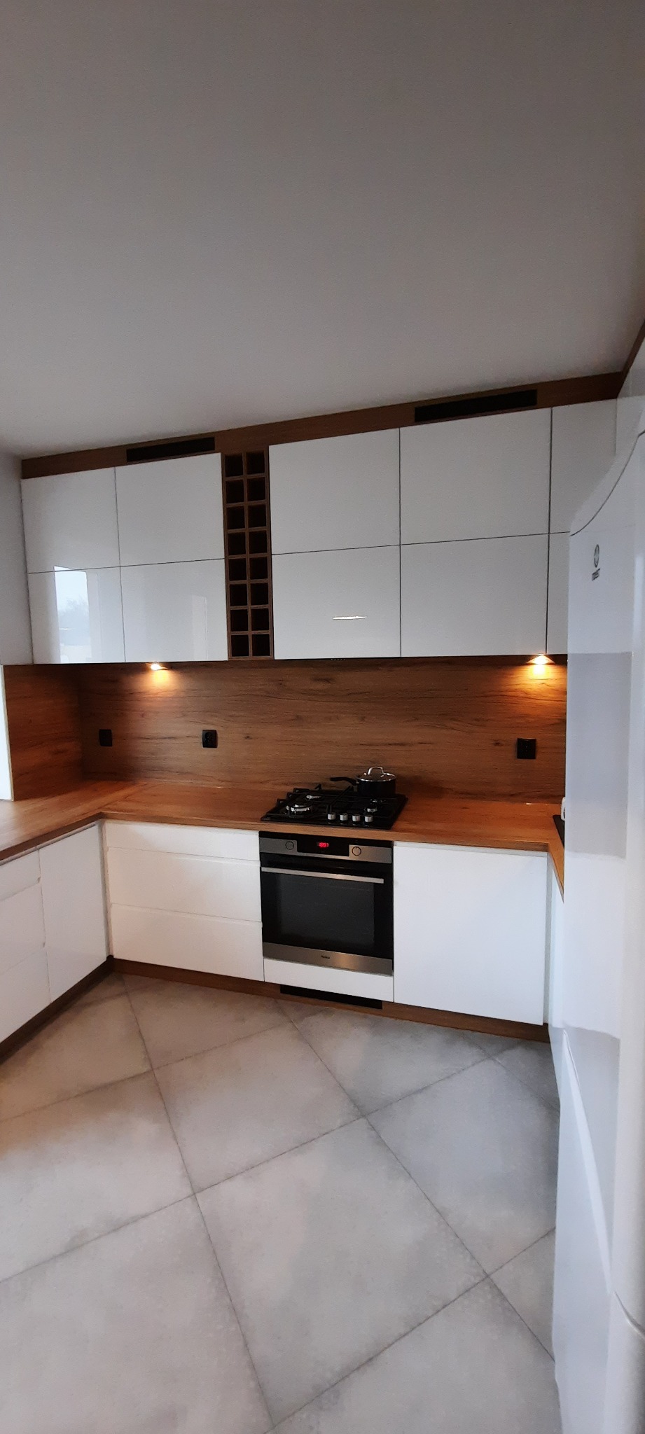 Nowoczesna kuchnia z białymi, lakierowanymi szafkami, drewnianym blatem i panelem ściennym, wbudowaną płytą grzewczą i piekarnikiem oraz półkami na wino.