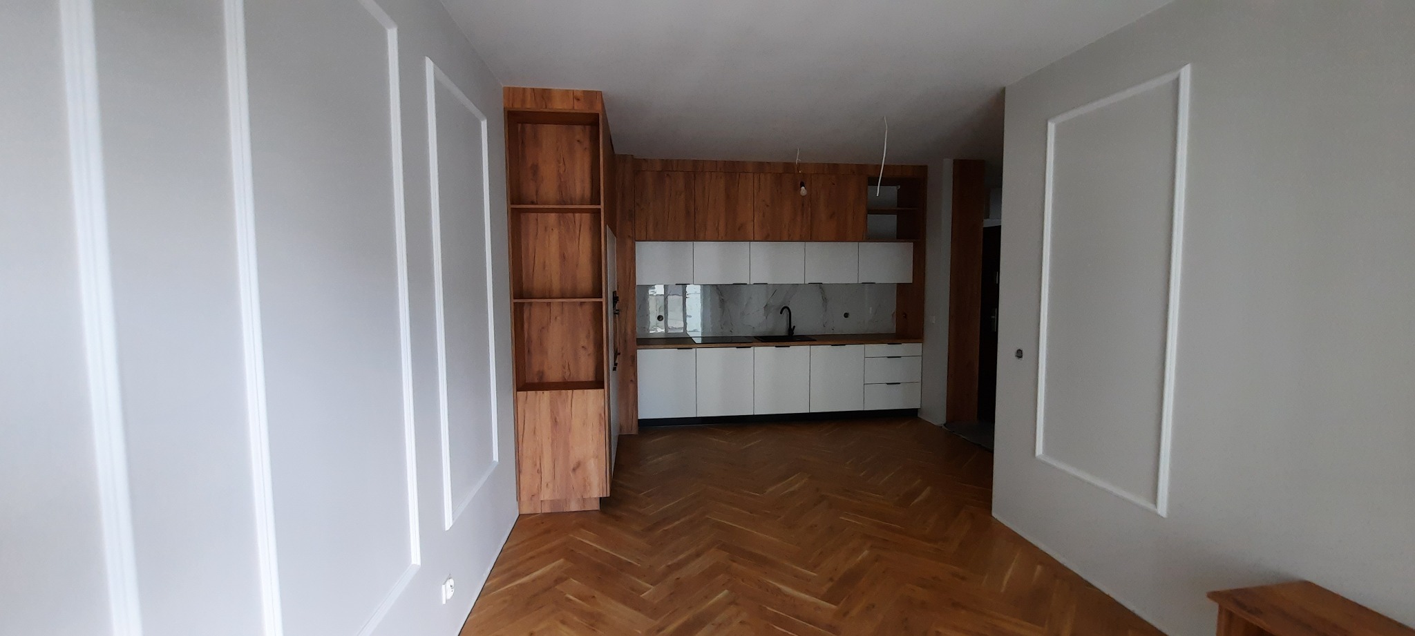 Nowoczesna kuchnia z białymi szafkami dolnymi i drewnopodobnymi górnymi, z czarną armaturą i marmurowym backsplash, widoczna z perspektywy szerokokątnej, z parkietem w jodełkę na podłodze...