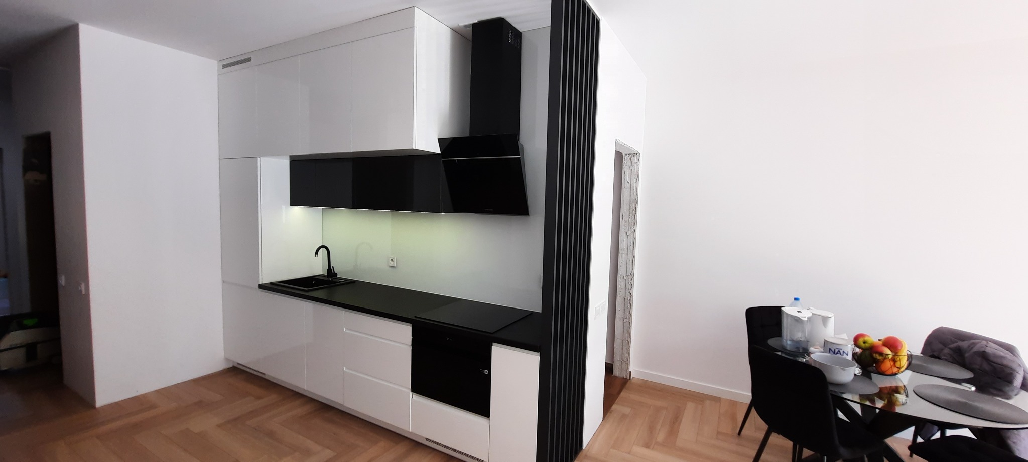 Nowoczesna kuchnia z białymi, lakierowanymi szafkami, czarnym blatem i okapem, widoczny fragment jadalni ze szklanym stołem i owocami.
