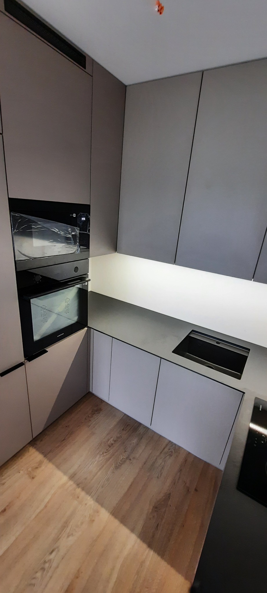 Nowoczesna kuchnia z szarymi frontami szafek, wbudowanym piekarnikiem Samsung w folii ochronnej i drewnianą podłogą.
