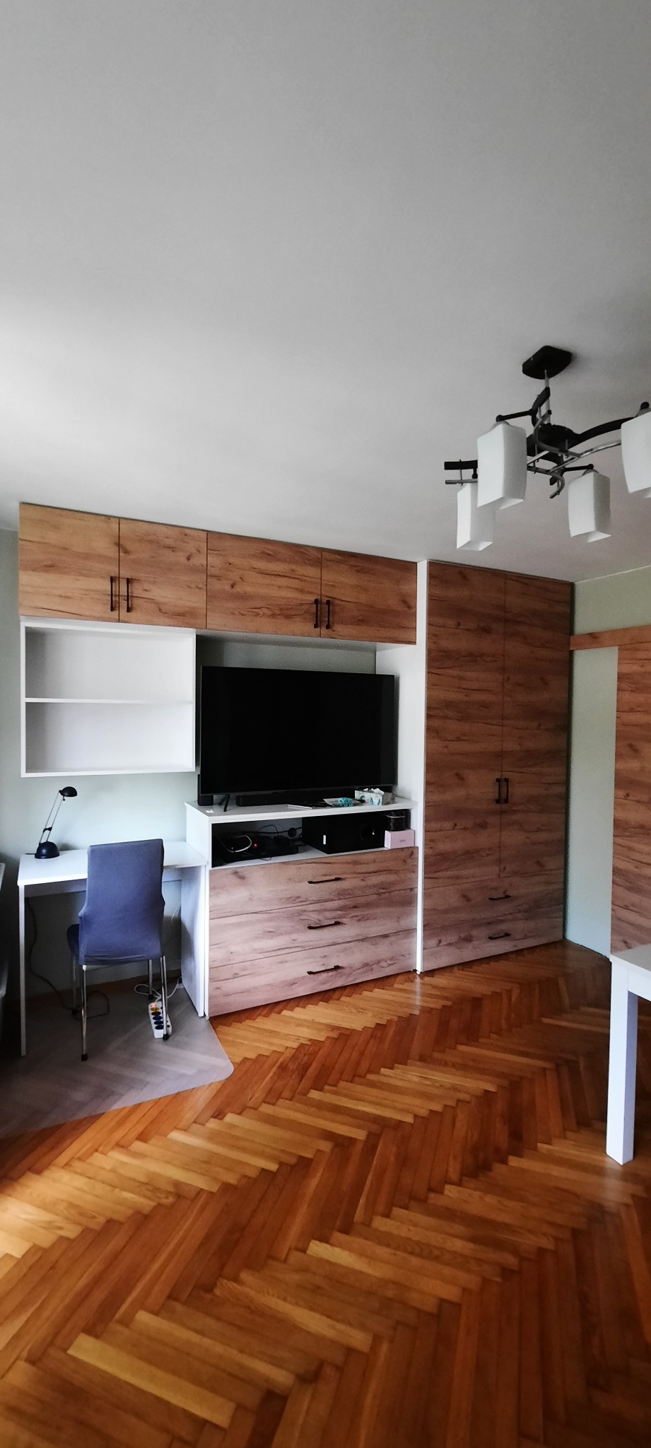 Zabudowa meblowa w pokoju z biurkiem, szafą i szafkami wiszącymi, wykończona okleiną drewnopodobną, na podłodze parkiet w jodełkę.