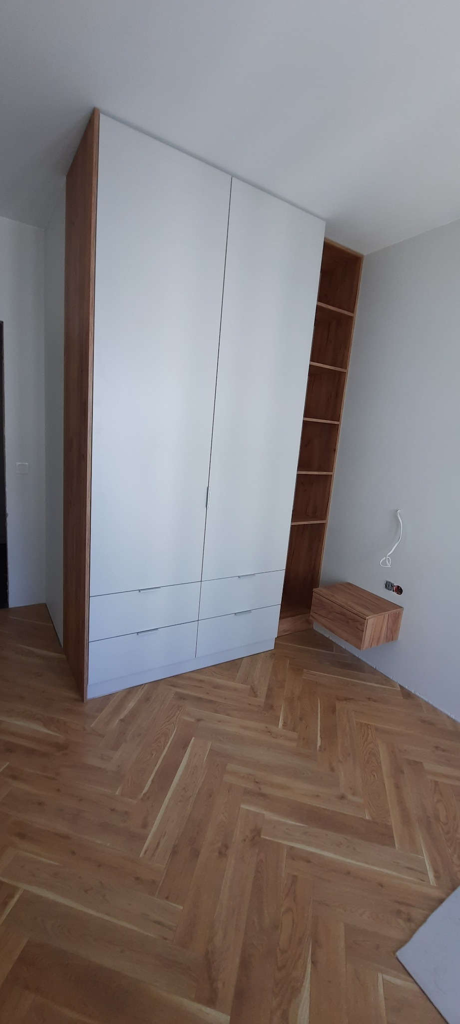 Biała szafa z drewnianymi bokami i szufladami, uzupełniona drewnianą półką wiszącą, na podłodze parkiet ułożony w jodełkę.