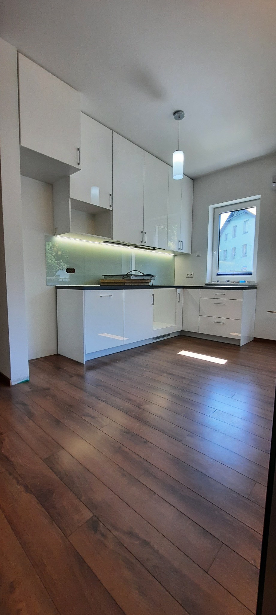 Nowoczesna, biała kuchnia z połyskiem, podświetlany panel szklany nad blatem, drewniana podłoga, widok z poziomu podłogi.