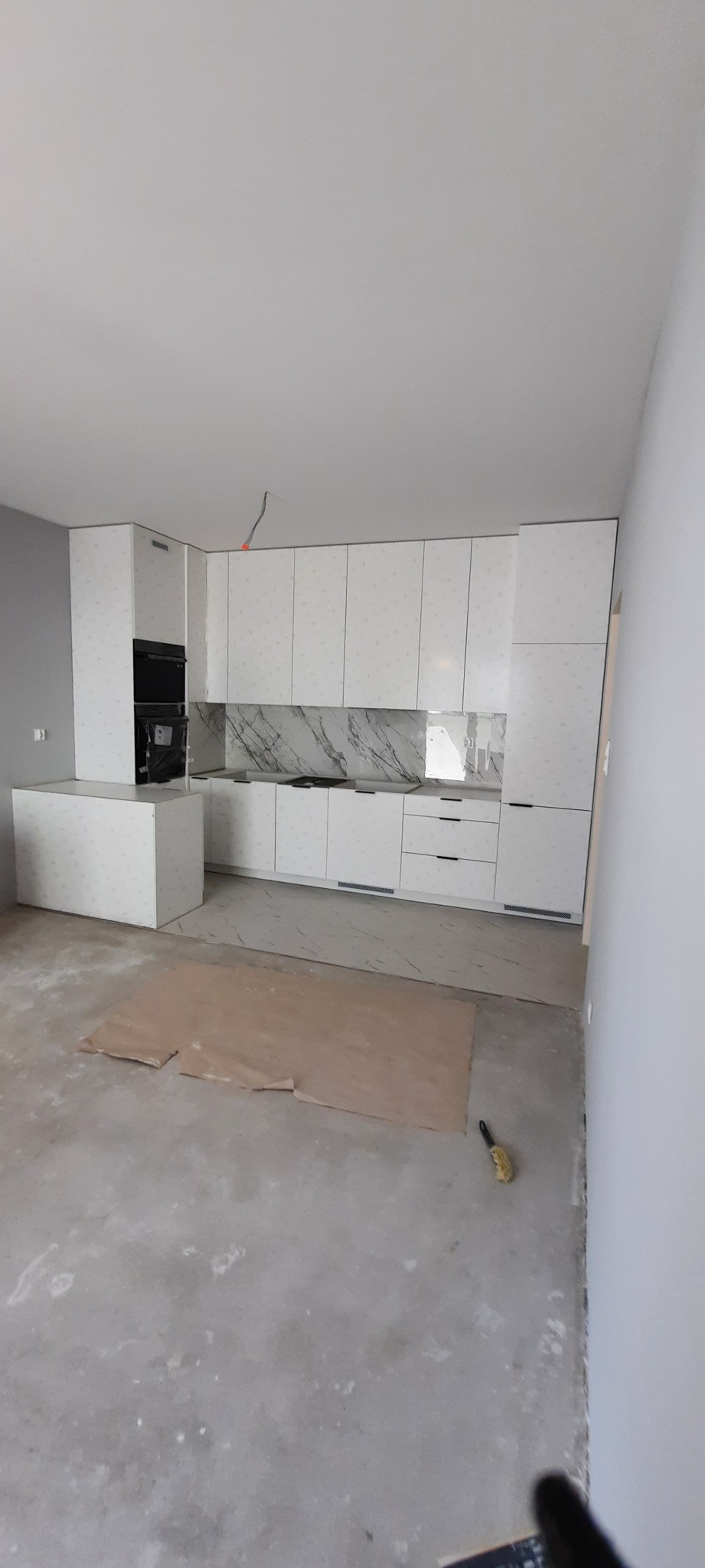 Jasna kuchnia z białymi frontami szafek z delikatnym wzorem, marmurowy backsplash, częściowo wykończone podłogi, widoczne zabezpieczenia podłogi i narzędzie malarskie.