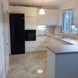 Jasna kuchnia z wyspą, fronty w kolorze złamanej bieli, czarny sprzęt AGD, płytki imitujące kamień na podłodze i ścianie nad blatem, nowoczesne oświetlenie.