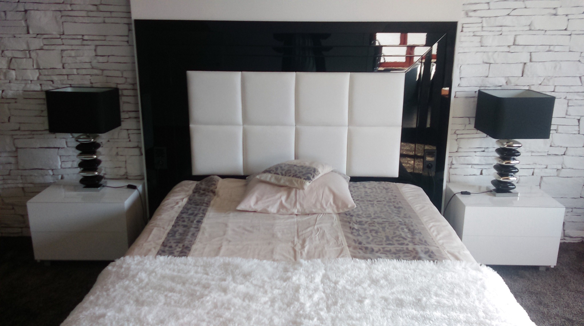 Nowoczesna sypialnia z łóżkiem z białym tapicerowanym zagłówkiem, czarną ramą, białymi szafkami nocnymi i lampkami z czarnymi abażurami na tle ściany z imitacją kamienia.