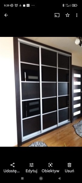 Ciemna szafa wnękowa z przesuwnymi drzwiami z czarnego szkła i srebrnymi ramami, widoczna z boku w pokoju z drewnianą podłogą i częściowo widocznymi drzwiami.