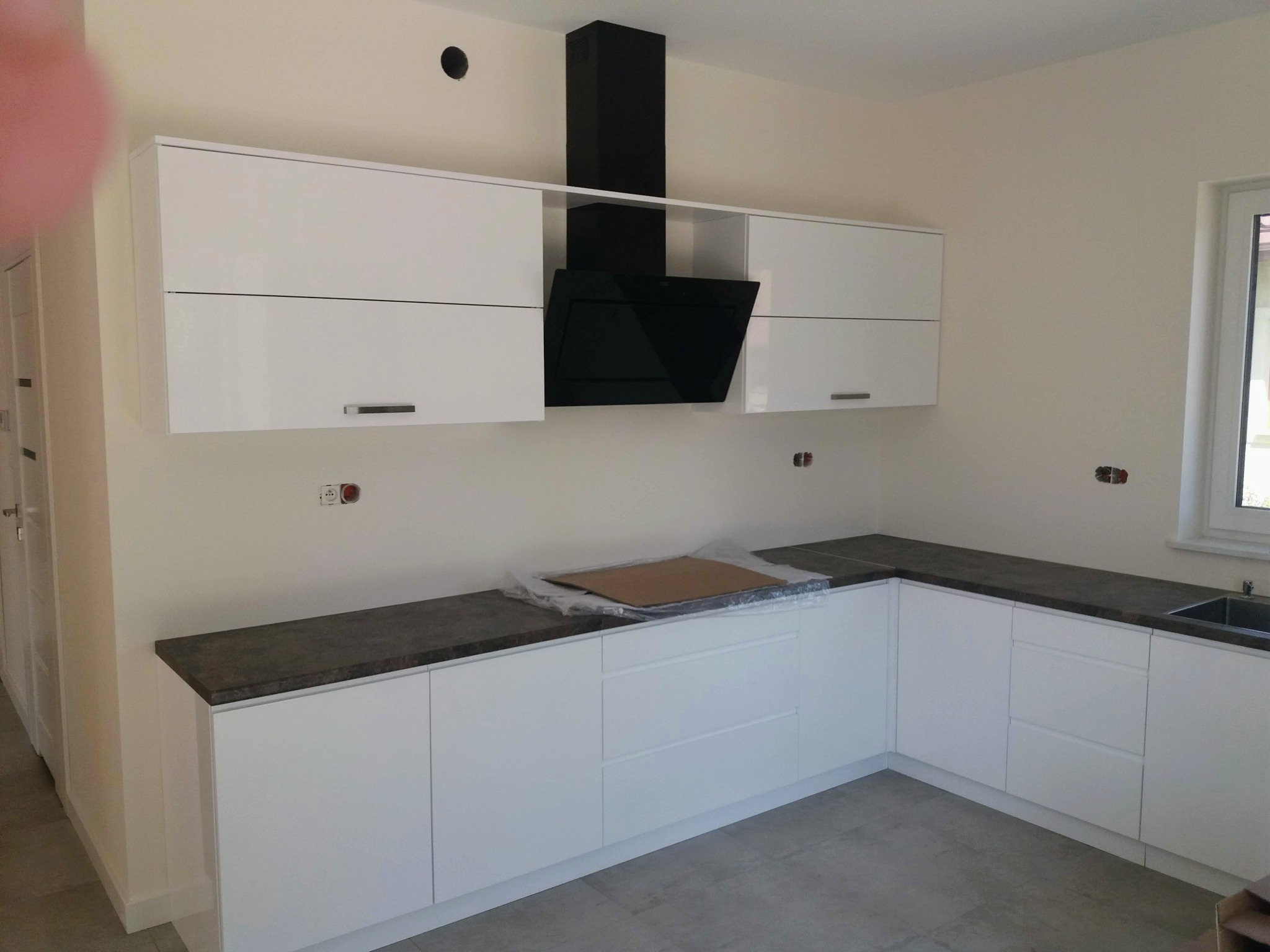 Nowoczesna, biała zabudowa kuchenna z ciemnym blatem i czarnym okapem, widoczne ślady po montażu na ścianie, karton zabezpieczający na blacie.