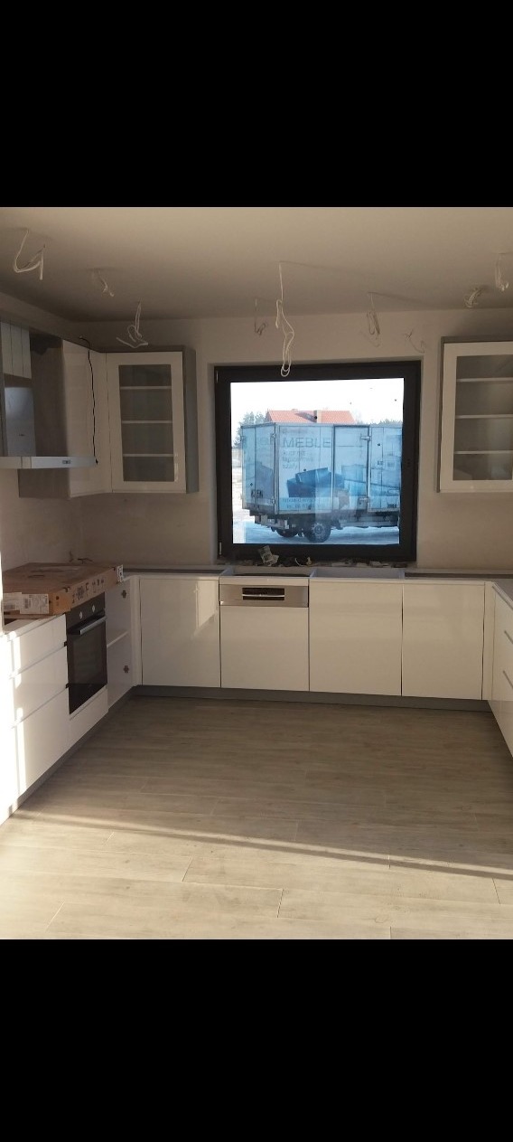 Wnętrze kuchni w trakcie montażu z widocznym przez okno samochodem dostawczym z napisem MEBLE, niedokończone podłączenia elektryczne zwisają z sufitu.