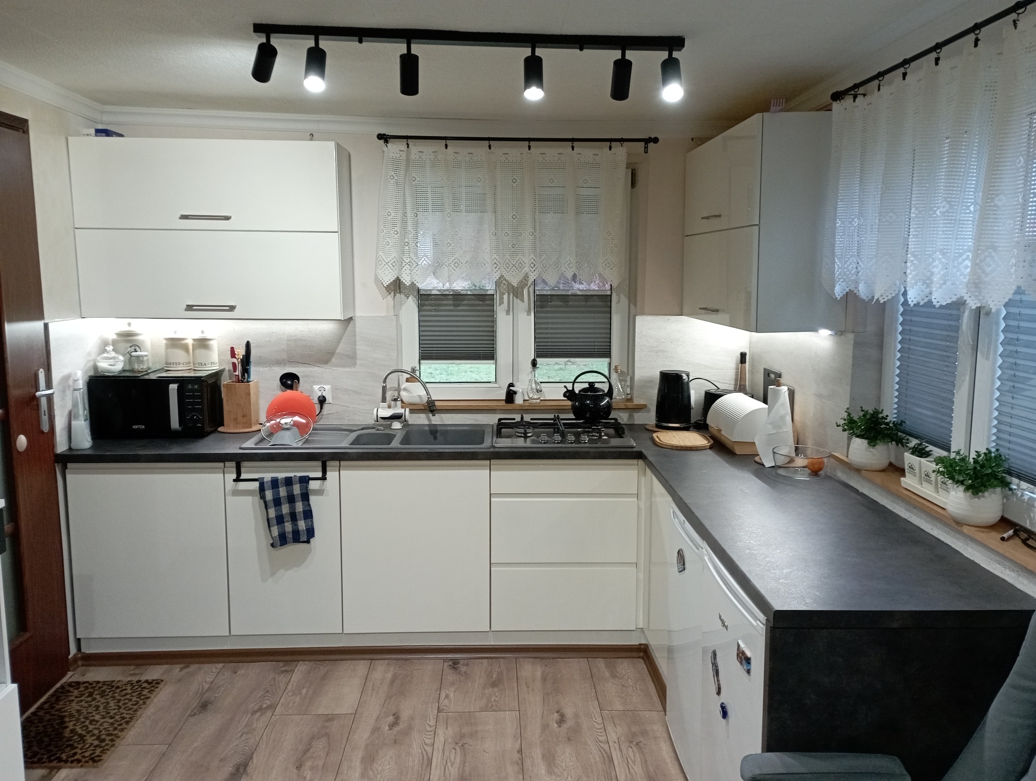 Jasna kuchnia z białymi frontami szafek, szarym blatem, drewnianą podłogą i oświetleniem punktowym. Okno zdobi firanka z koronki i rolety plisowane.