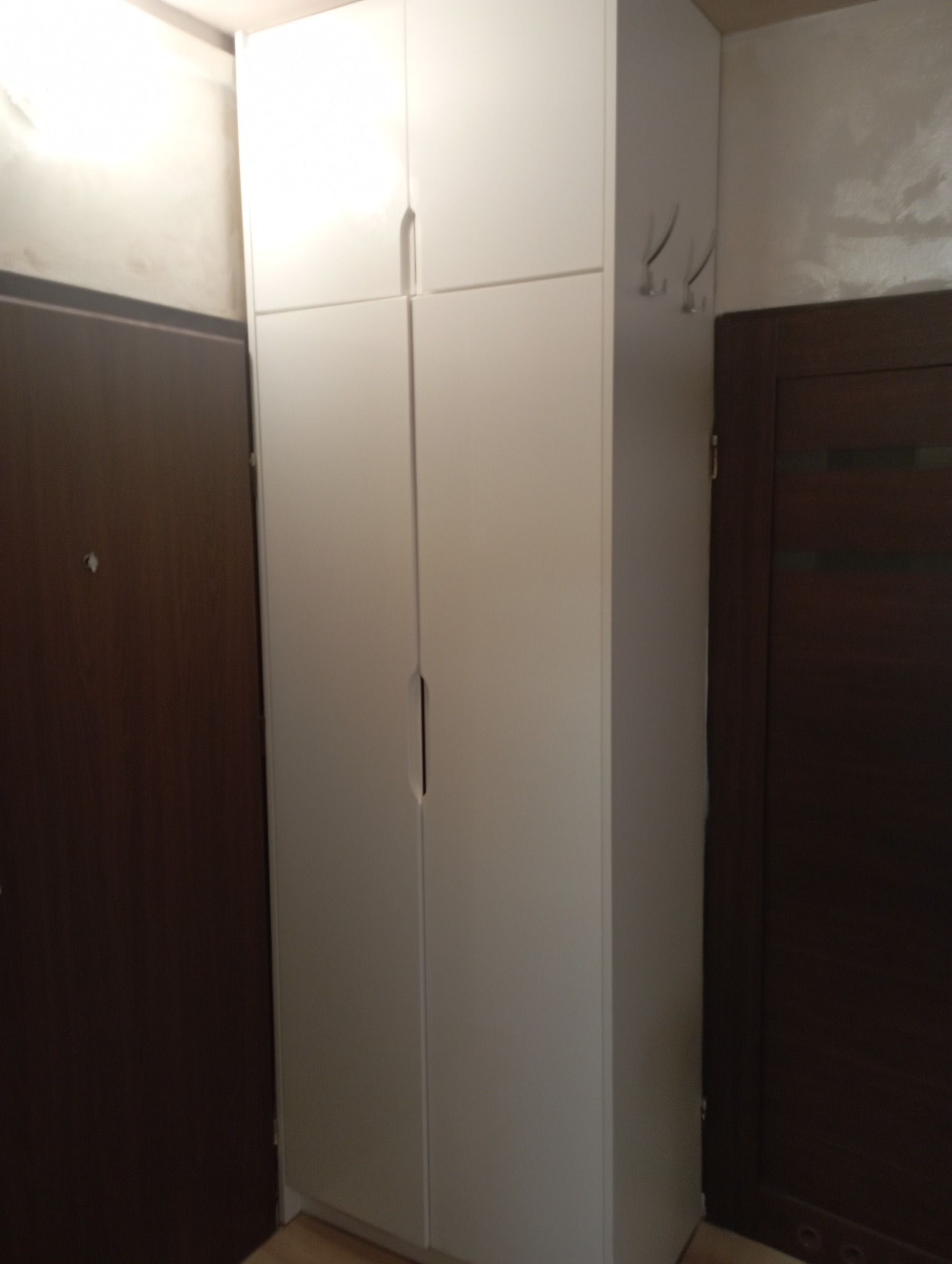 Wysoka, biała szafa z czterema drzwiami, wbudowana we wnękę między dwoma drzwiami w mieszkaniu. Po prawej stronie szafy widoczne haczyki na ścianie.