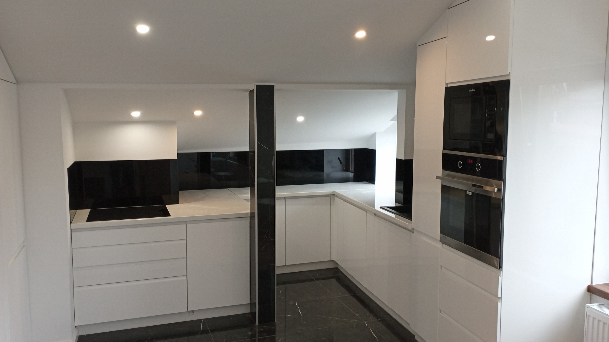 Biała, nowoczesna kuchnia z czarnymi akcentami, błyszczące fronty szafek, czarny panel nad blatem, podłoga z czarnego marmuru, oświetlenie punktowe.