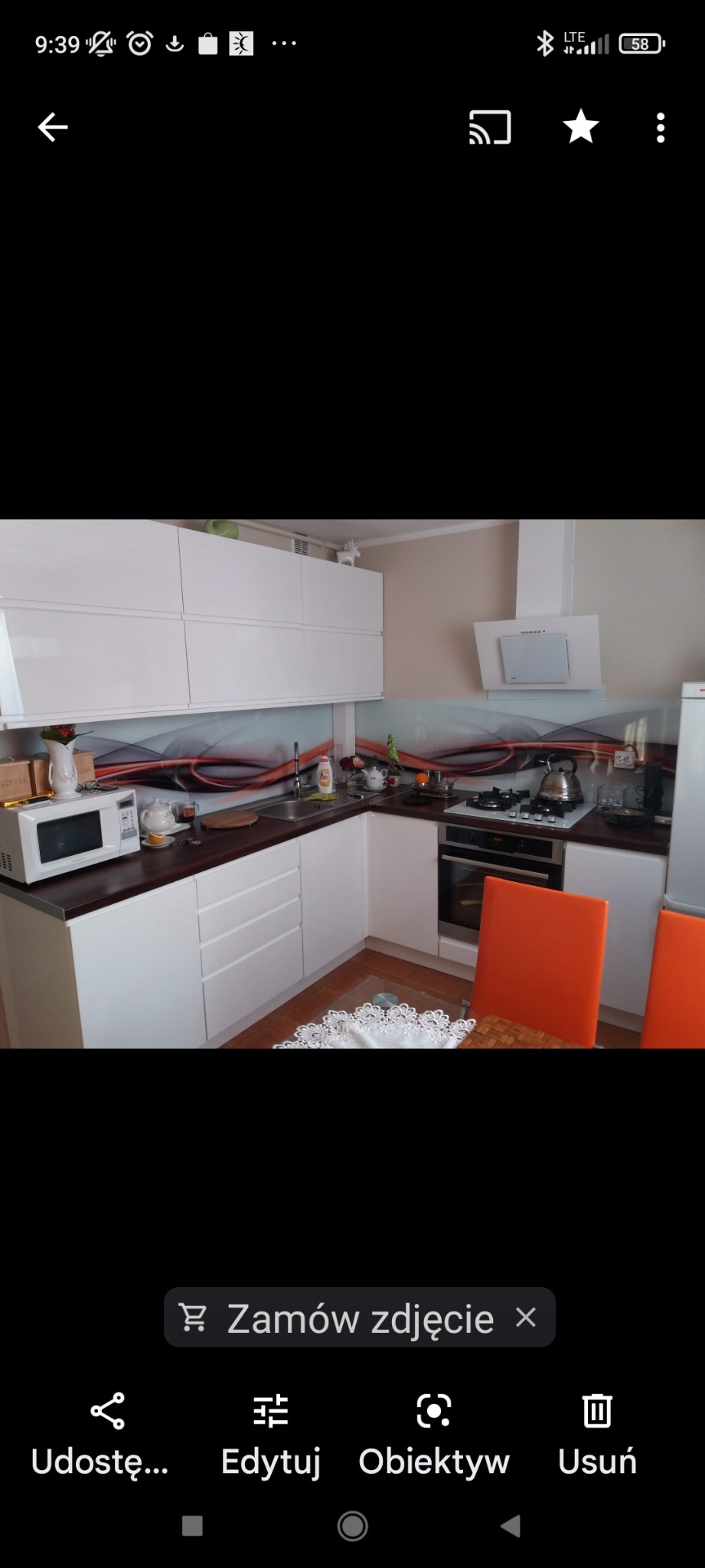 Jasna kuchnia z brązowymi blatami, szklanym panelem z abstrakcyjnym wzorem, widoczne pomarańczowe krzesła i sprzęt AGD.