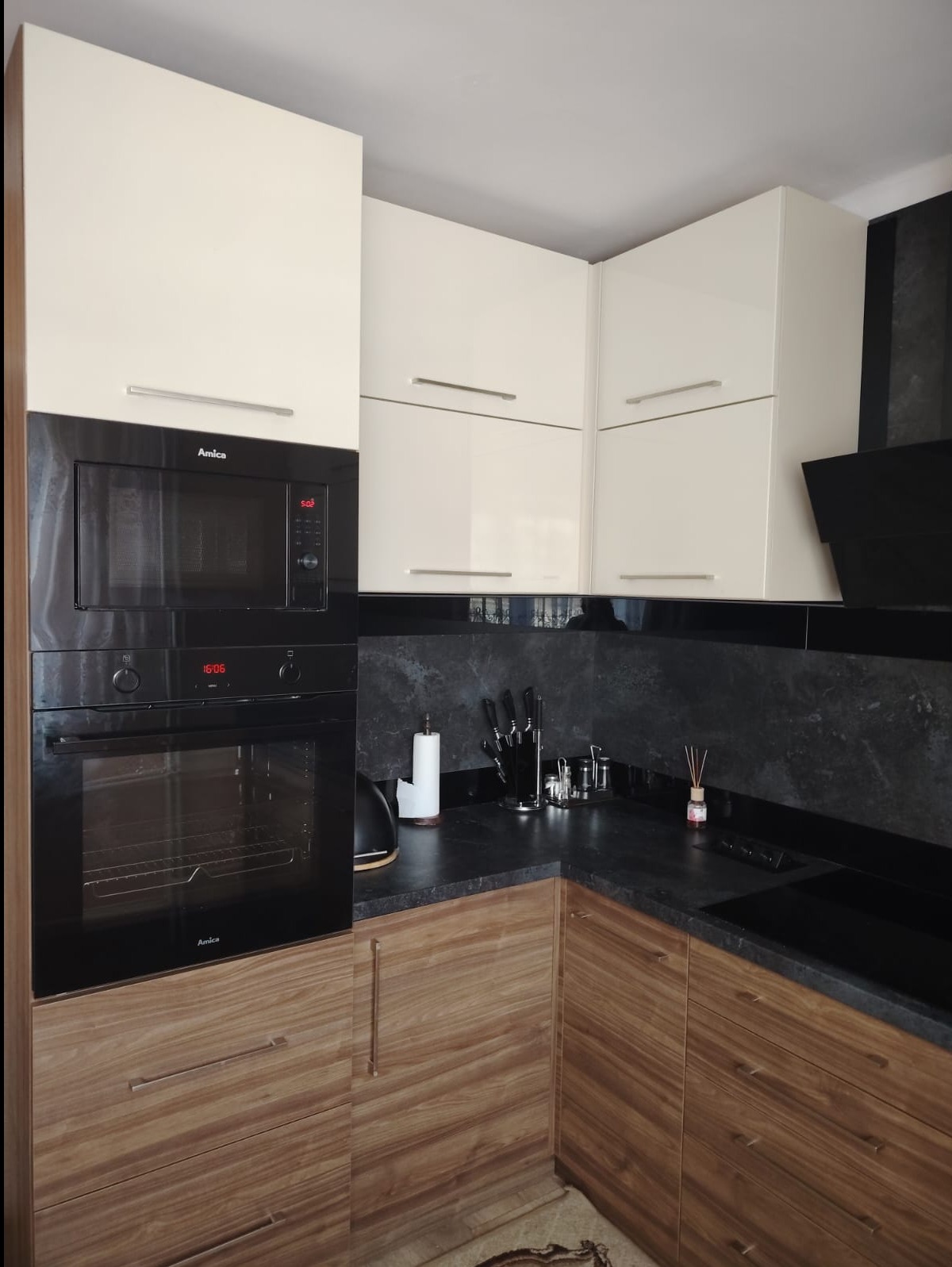 Nowoczesna kuchnia z drewnianymi szafkami dolnymi, białymi szafkami górnymi i czarnym blatem z kamienia. Widoczny sprzęt AGD marki Amica: piekarnik i mikrofalówka. Na blacie stojak z nożami...