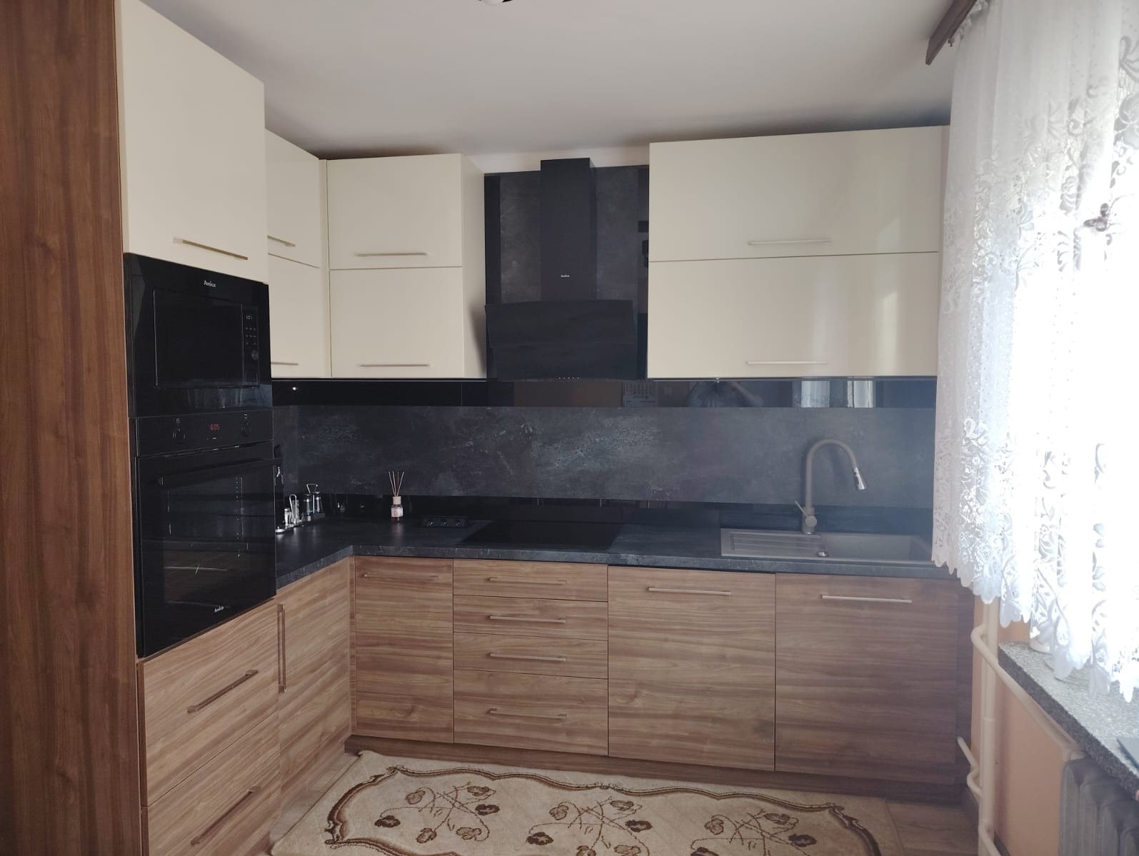 Nowoczesna kuchnia z zabudową w odcieniach beżu i drewna, z czarnym panelem nad blatem i piekarnikiem wbudowanym w wysoką szafkę. Na podłodze dywan z orientalnym wzorem.