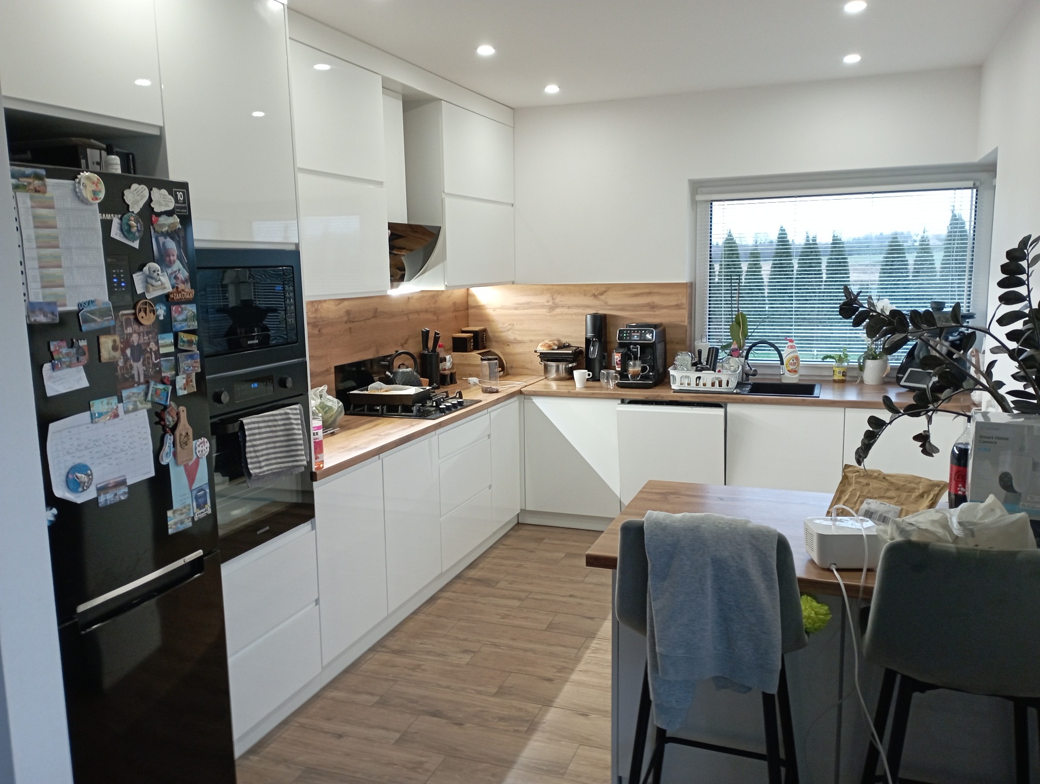 Nowoczesna kuchnia z białymi szafkami, drewnianym blatem i czarną lodówką ozdobioną magnesami, widok na okno z żaluzjami i rzędem drzew.