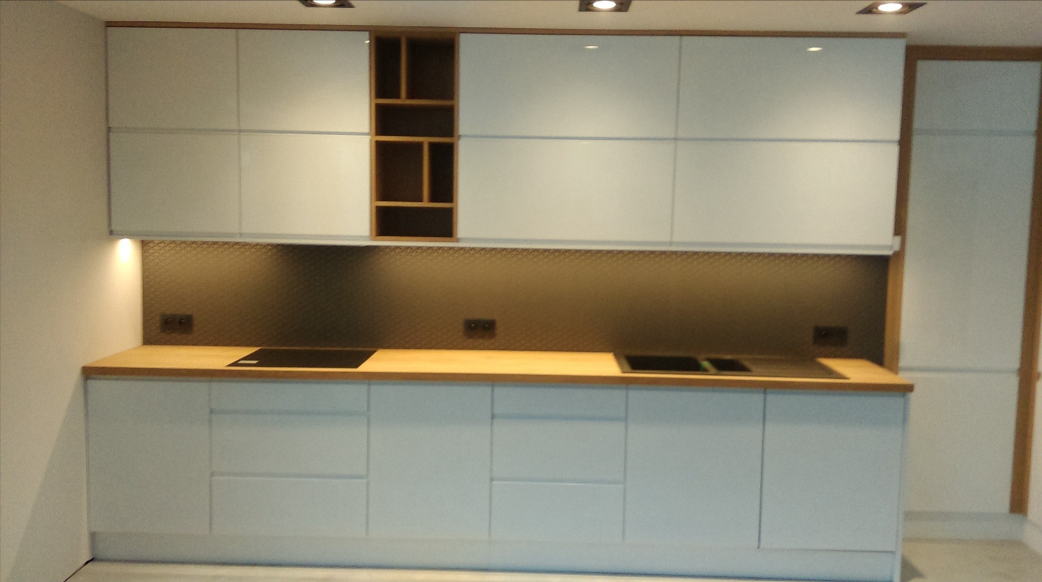 Nowoczesna kuchnia z białymi frontami szafek, drewnianym blatem i otwartą półką z drewnianymi przegródkami, widoczne gniazdka elektryczne na ścianie z mozaikową okładziną.