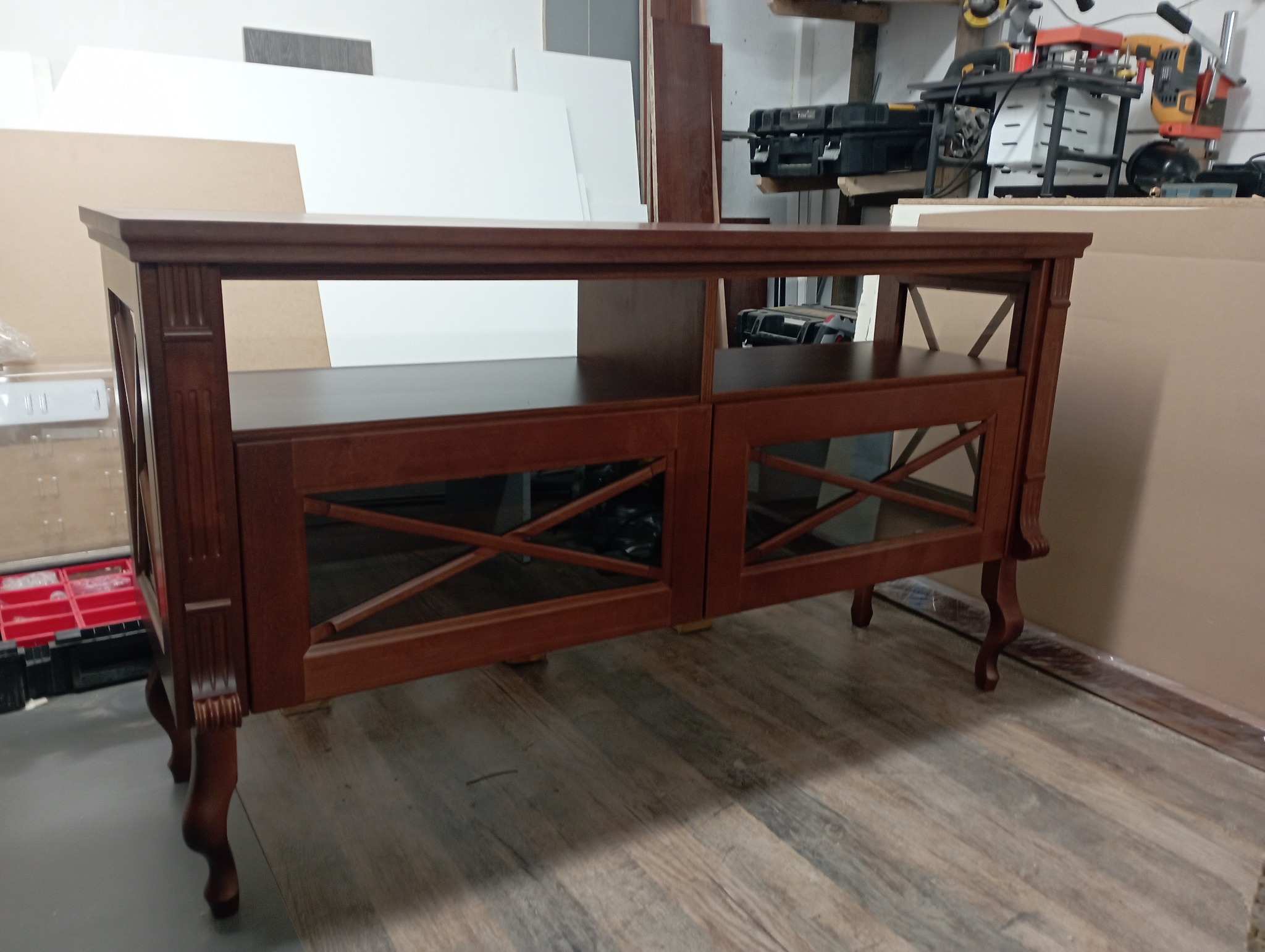 Ręcznie wykonana, drewniana szafka w stylu retro z dekoracyjnymi nogami i zdobieniami, stojąca w warsztacie na podłodze z paneli. Widoczne narzędzia w tle.