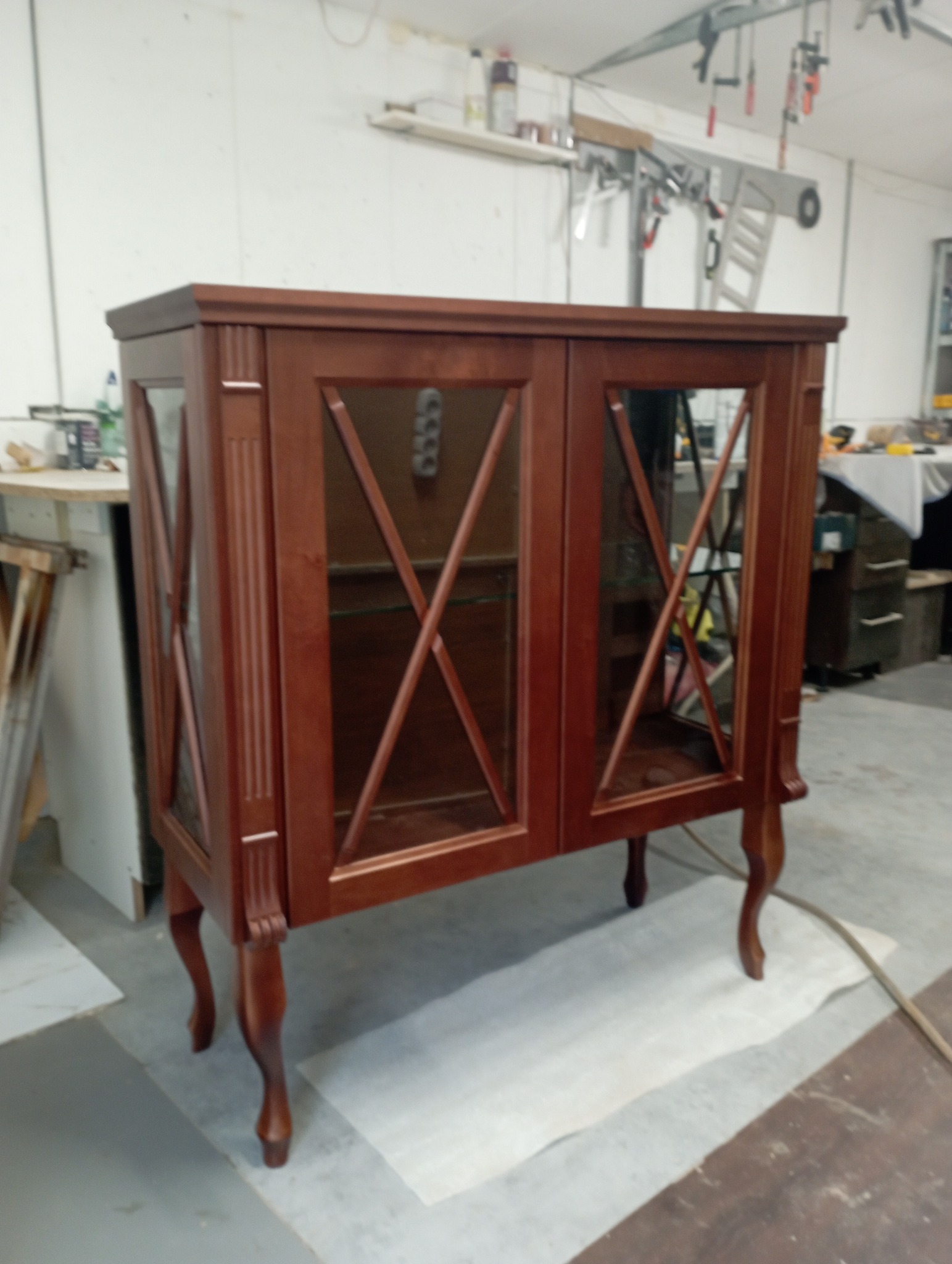 Ręcznie robiona, mahoniowa witryna z dwoma przeszklonymi drzwiczkami, osadzona na giętych nóżkach, stoi na podłodze w warsztacie.