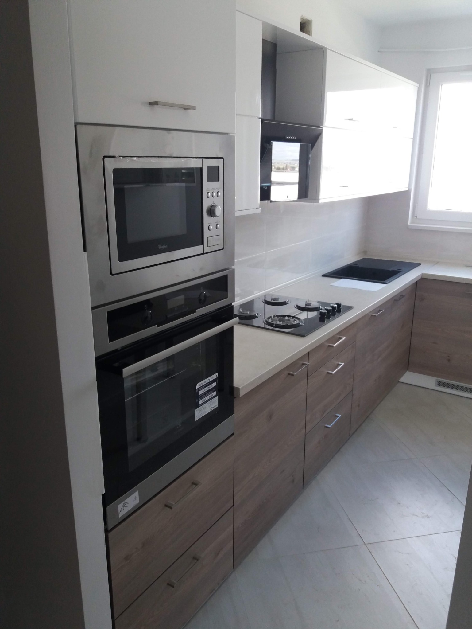 Nowoczesna kuchnia: białe szafki górne, dolne z forniru drewna, zabudowany piekarnik i mikrofalówka Whirlpool, czarna płyta gazowa, jasny blat i podłoga.