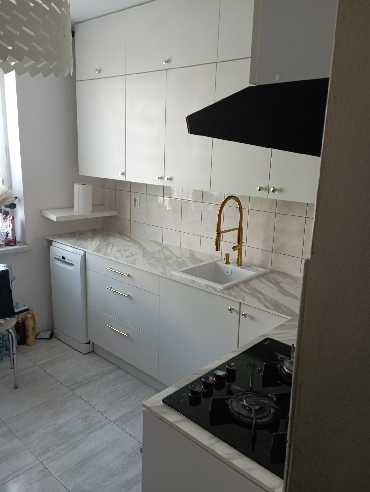 Jasna kuchnia z białymi szafkami i złotą armaturą. Blat imitujący marmur i czarna płyta grzewcza dodają elegancji.