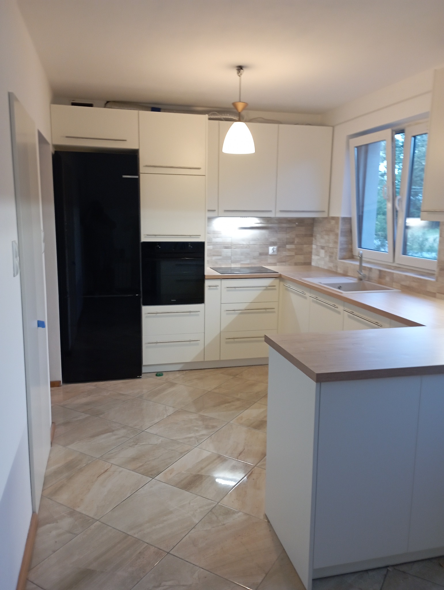 Jasna kuchnia z wyspą, fronty w kolorze złamanej bieli, czarny sprzęt AGD, płytki imitujące kamień na podłodze i ścianie nad blatem, nowoczesne oświetlenie.