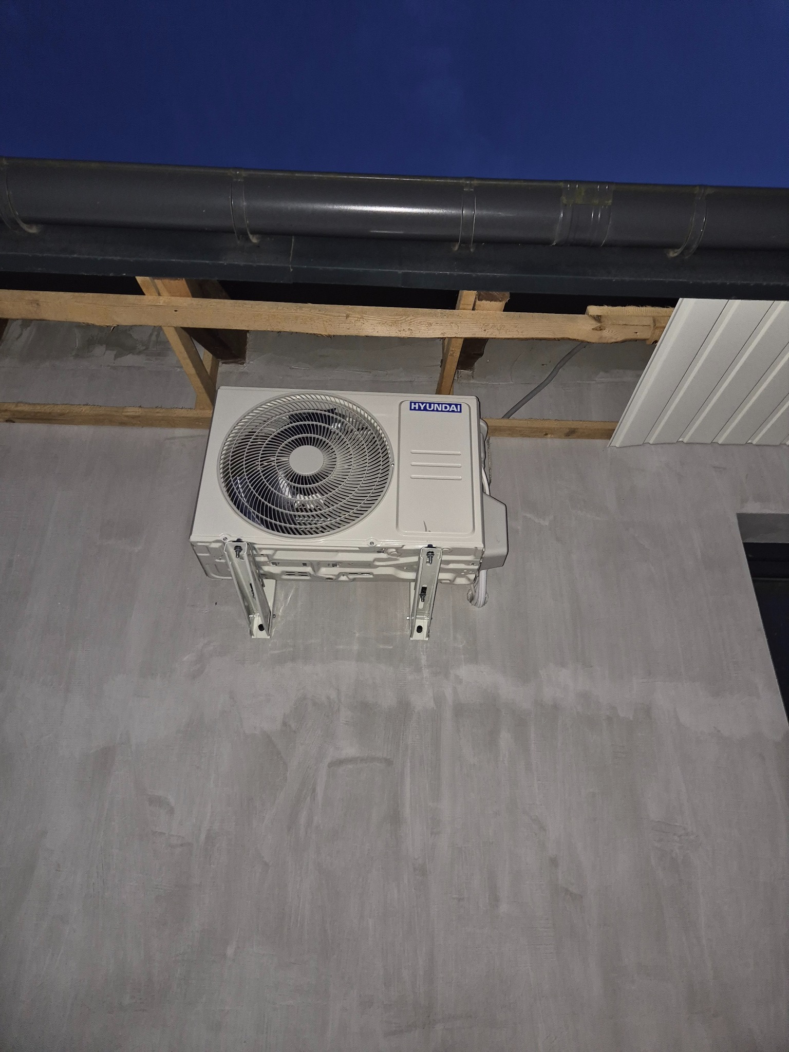 Zamontowana jednostka zewnętrzna klimatyzacji Hyundai na szarej ścianie pod zadaszeniem. Widoczne elementy montażowe i instalacja elektryczna. Nocne ujęcie.