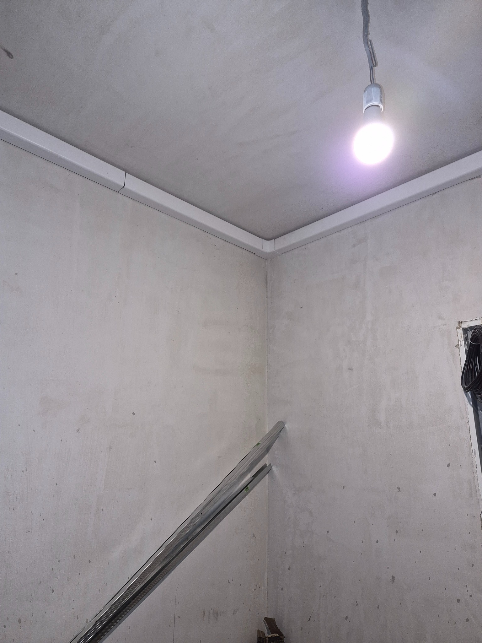 Montaż białych listew maskujących na suficie w rogu pomieszczenia. Widoczna surowa ściana i wisząca żarówka. Elementy instalacji wentylacyjnej w trakcie montażu.