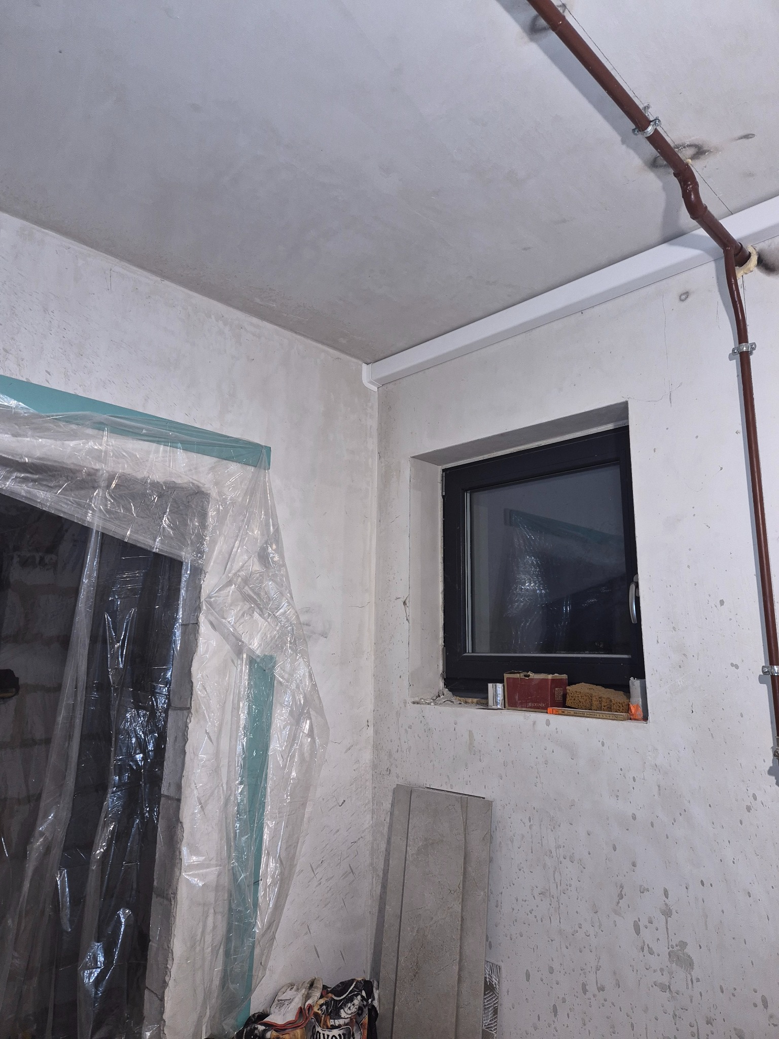 Rura instalacji pod sufitem w surowym wnętrzu, zabezpieczone folią drzwi, okno z czarną ramą. Ściany w trakcie prac wykończeniowych, widoczne ślady po zaprawie.