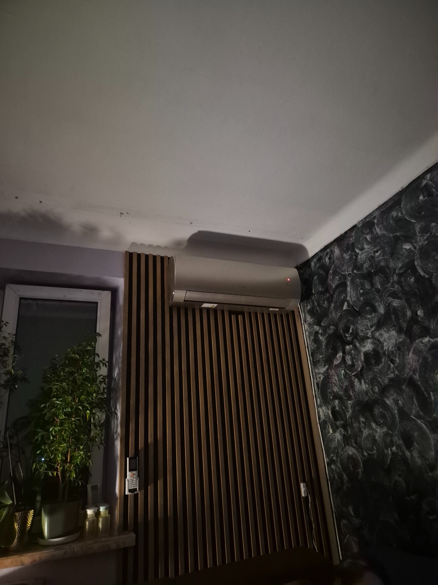 Nowoczesny klimatyzator ścienny w jasnym wnętrzu, z dekoracyjną ścianką z drewnianych listew i wzorzystą tapetą. Widoczny pilot i roślina doniczkowa.