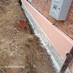 GAWEŁCZYK IZOLACJE NATRYSKOWE - Ocieplenie fundamentów pianką PUR i styropianem XPS przy ceglanym budynku z przyłączem gazowym. Widoczny wykop i rura kanalizacyjna.