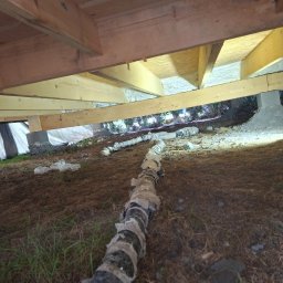 GAWEŁCZYK IZOLACJE NATRYSKOWE - Niskie ujęcie spod drewnianej konstrukcji podłogi, ukazujące świeżo zaaplikowaną białą izolację piankową między belkami. Na ziemi widoczna owinięta rura i fragmenty folii.