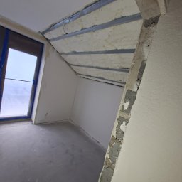 GAWEŁCZYK IZOLACJE NATRYSKOWE - Wnętrze poddasza z oknem zabezpieczonym niebieską folią, ukazujące natryskową izolację piankową między metalowymi profilami na skośnym suficie, obok surowej betonowej belki.