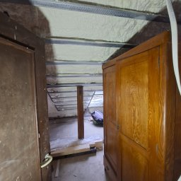 GAWEŁCZYK IZOLACJE NATRYSKOWE - Widok na poddasze z jasną pianką izolacyjną między metalowymi profilami konstrukcji dachu. W kadrze częściowo widoczne drewniane drzwi i stara szafa.