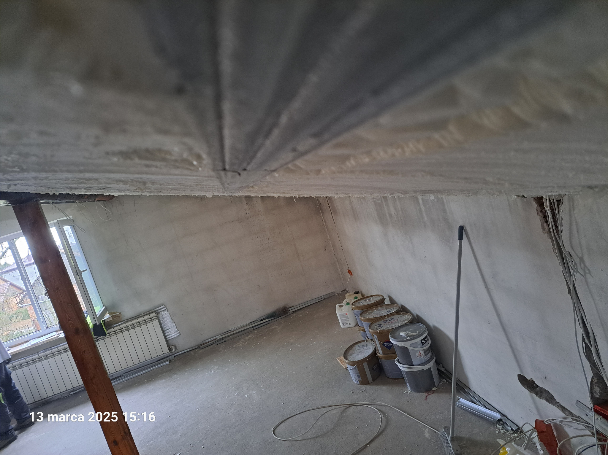 Sufit poddasza po natrysku pianki izolacyjnej, widoczne elementy konstrukcyjne, na podłodze wiadra z farbą i materiały budowlane, okno w tle.