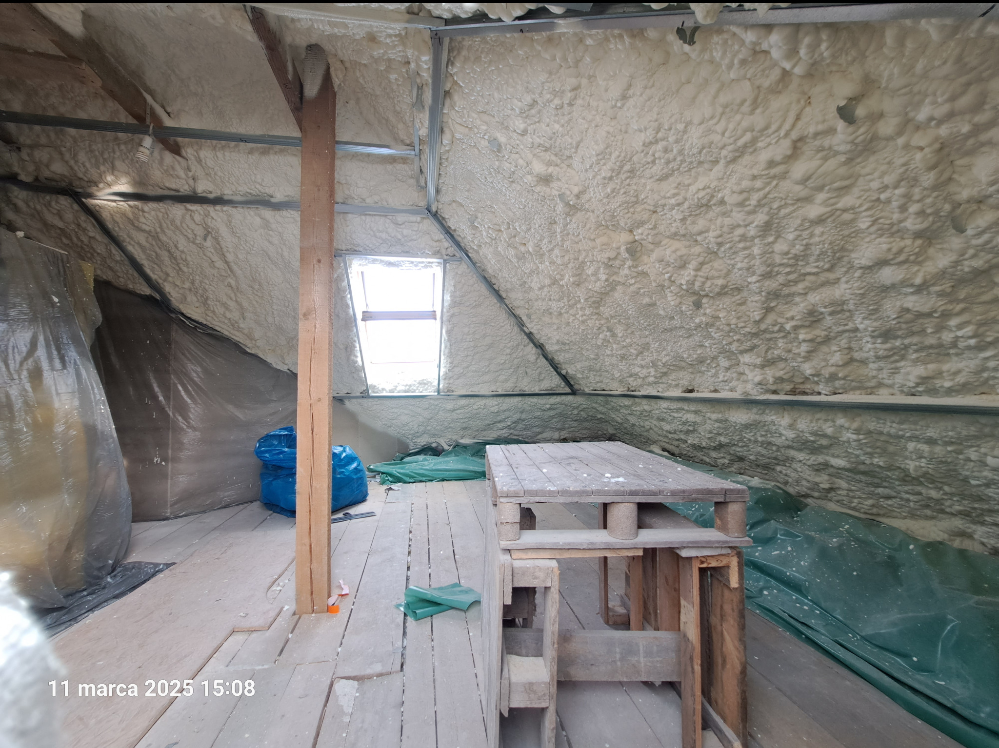 Poddasze z widoczną izolacją natryskową z pianki, drewnianą konstrukcją, oknem dachowym i prowizorycznym stołem z palet na drewnianej podłodze.