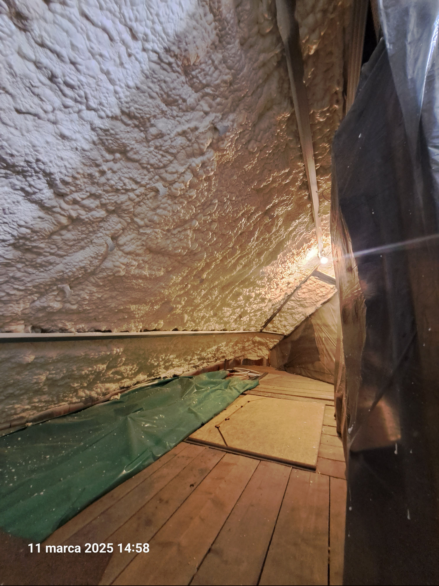 Poddasze z drewnianą podłogą, częściowo pokryte folią ochronną, po aplikacji izolacji natryskowej, widoczne nierównomierne pokrycie pianą.