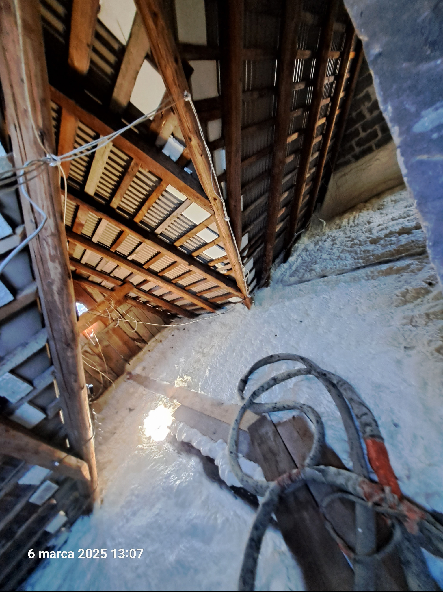 Poddasze z drewnianą konstrukcją pokryte warstwą białej pianki izolacyjnej, widoczne fragmenty więźby dachowej i przewody elektryczne.