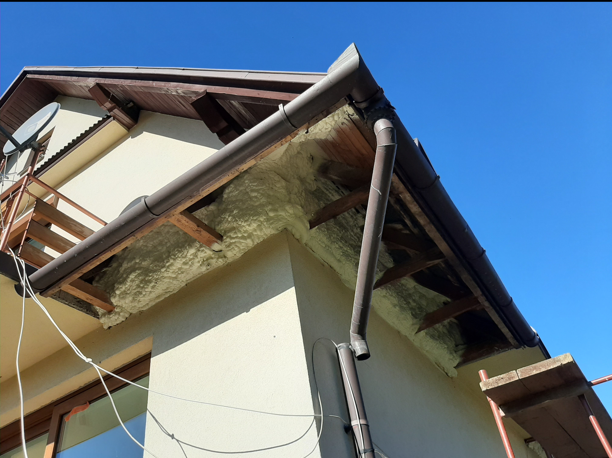 Fragment elewacji budynku z widoczną warstwą izolacji natryskowej w przestrzeni pod okapem, brązowe rynny i drewniana konstrukcja dachu na tle błękitnego nieba.