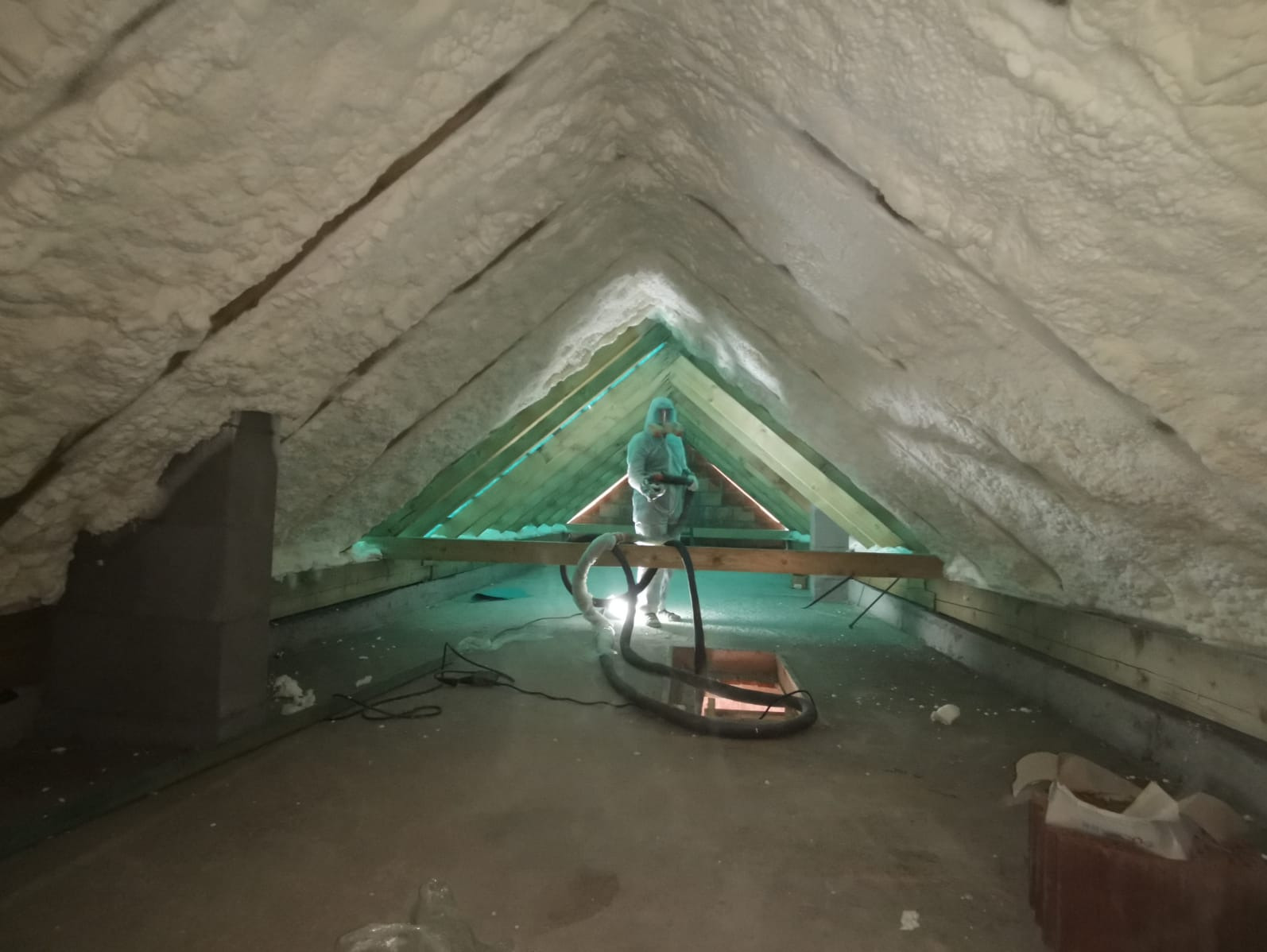 Pracownik w kombinezonie ochronnym aplikuje piankę izolacyjną na skośny dach poddasza, widoczne drewniane krokwie i instalacja do natrysku.