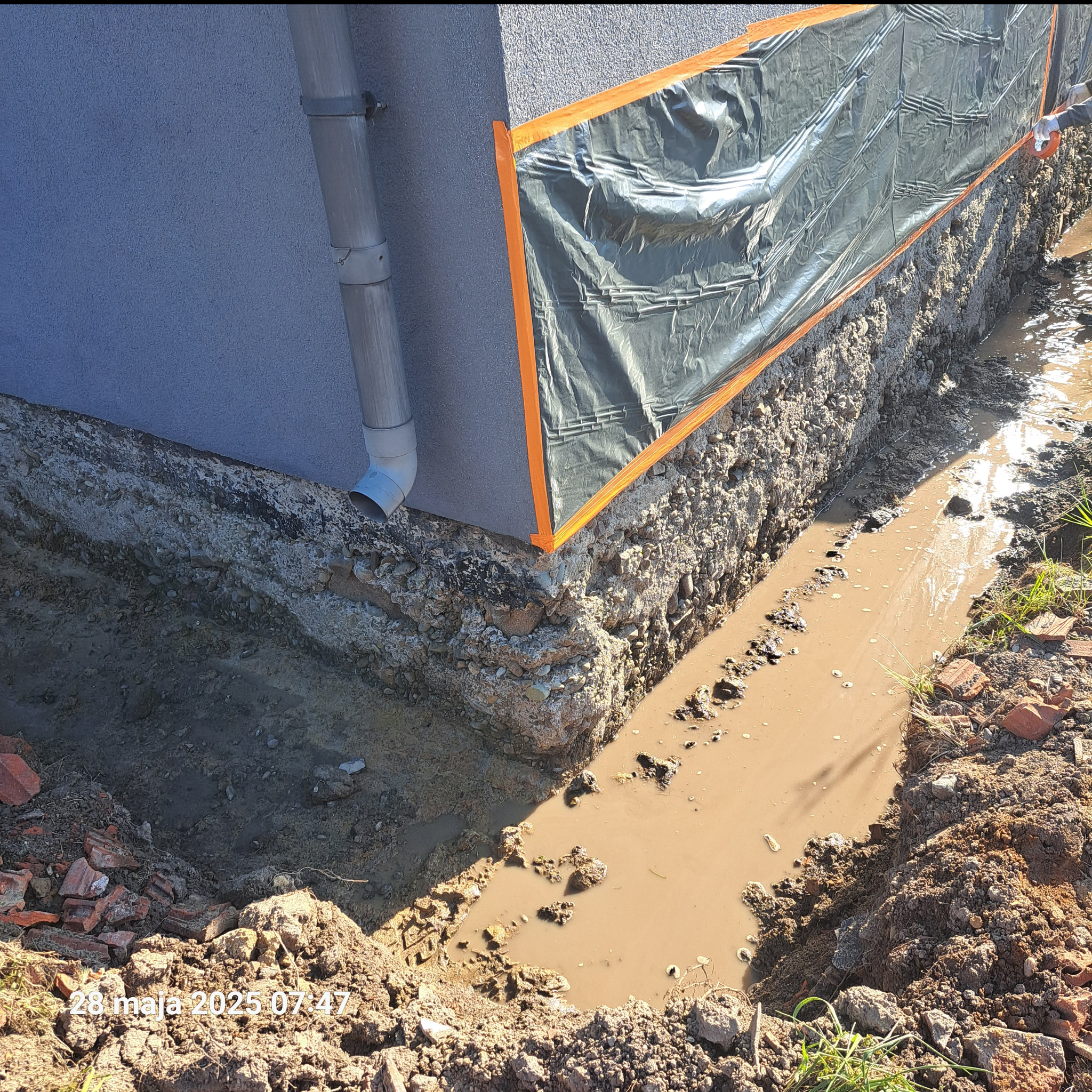 Fundament budynku zabezpieczony folią z pomarańczową taśmą, wykop zalany wodą. Widoczna rynna i fragment ściany budynku. Prace izolacyjne w toku.