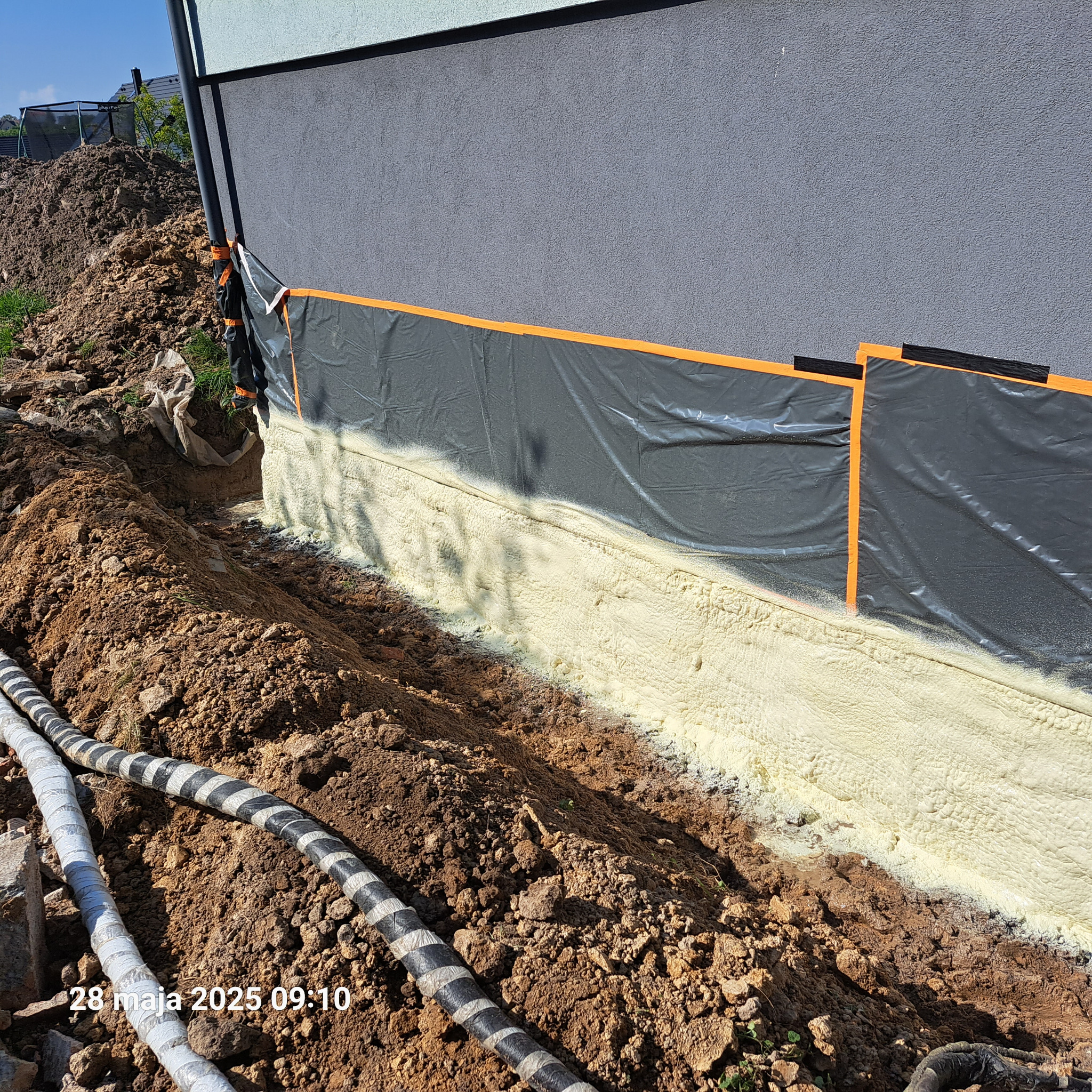 Fundament zabezpieczony pianką PUR, widoczne czarne folie ochronne z pomarańczowymi paskami i przewody. Ziemia wokół fundamentu.