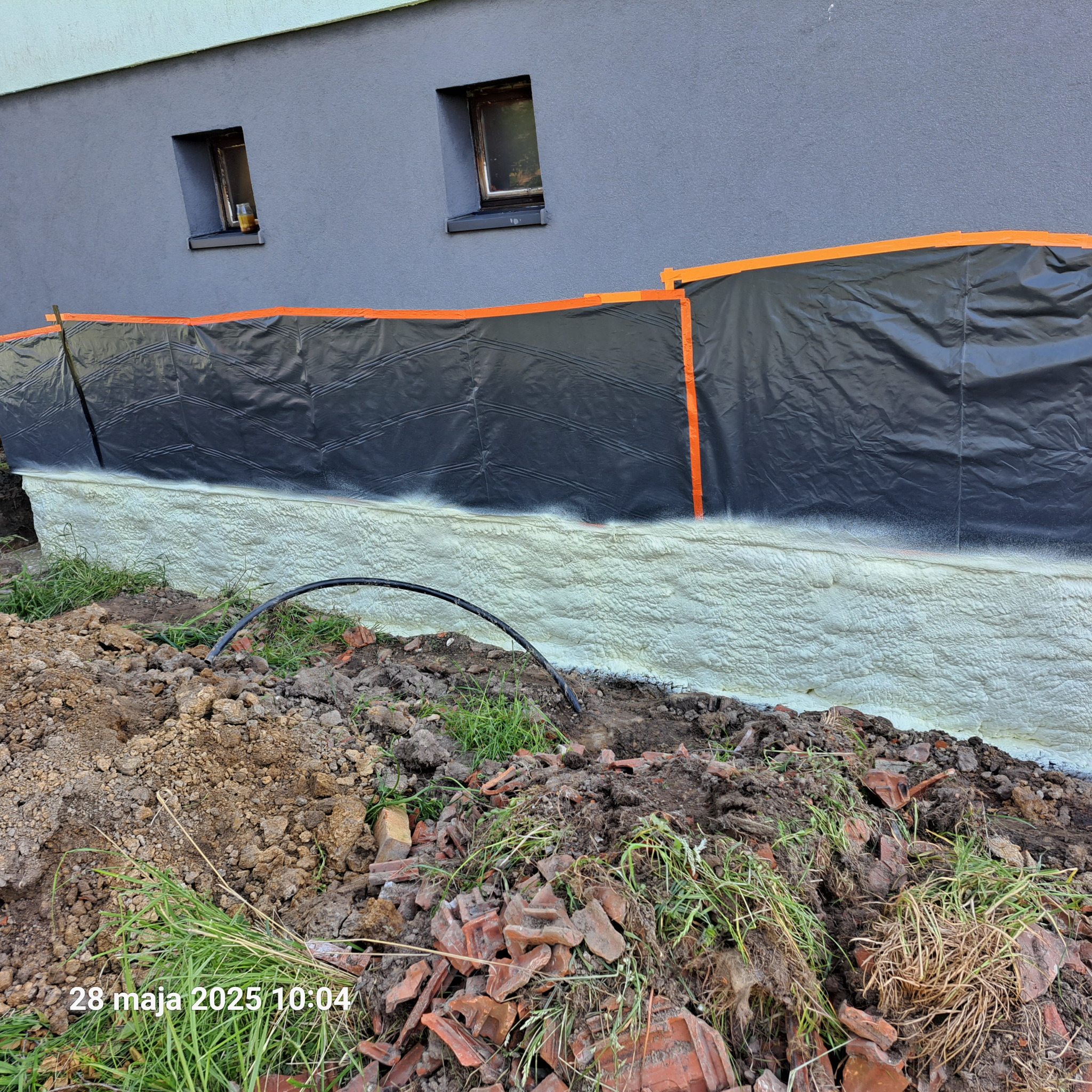 Ocieplenie fundamentów pianką PUR w trakcie realizacji. Widoczna czarna folia zabezpieczająca i świeżo nałożona pianka. W tle szara ściana budynku z oknami.